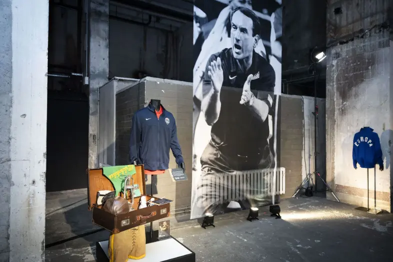 Présentation FIBA Hall of Fame BINK Un espace d'exposition sportive comprenant un buste BINK habillé d'une veste de basket USA à côté d'une valise de voyage vintage remplie d'équipements rétro lors de l'événement FIBA.