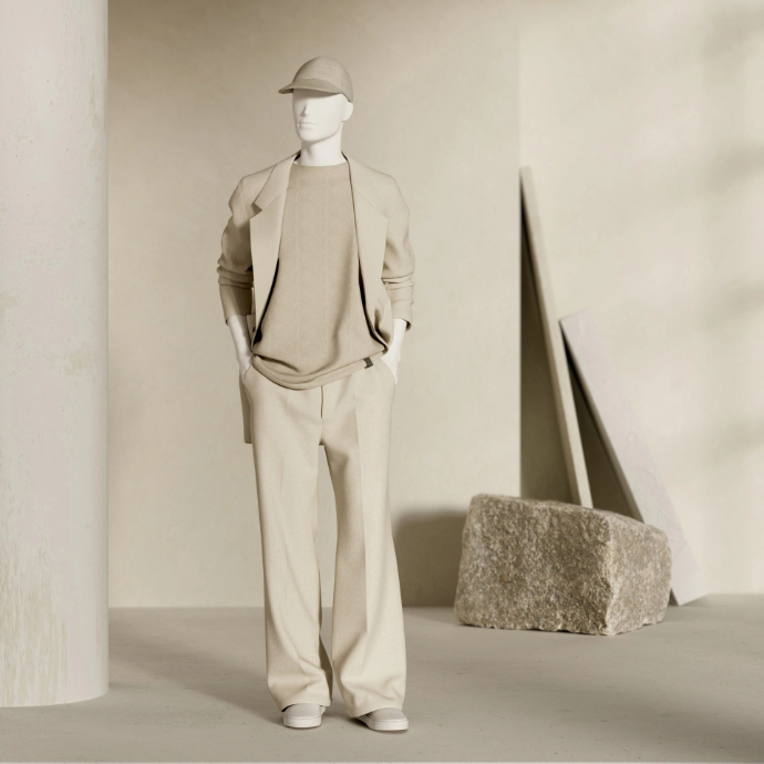 Male mannequin in a tonal beige outfit with a cap, jacket, knit top and wide trousers, standing next to a stone block in a sand-coloured setting