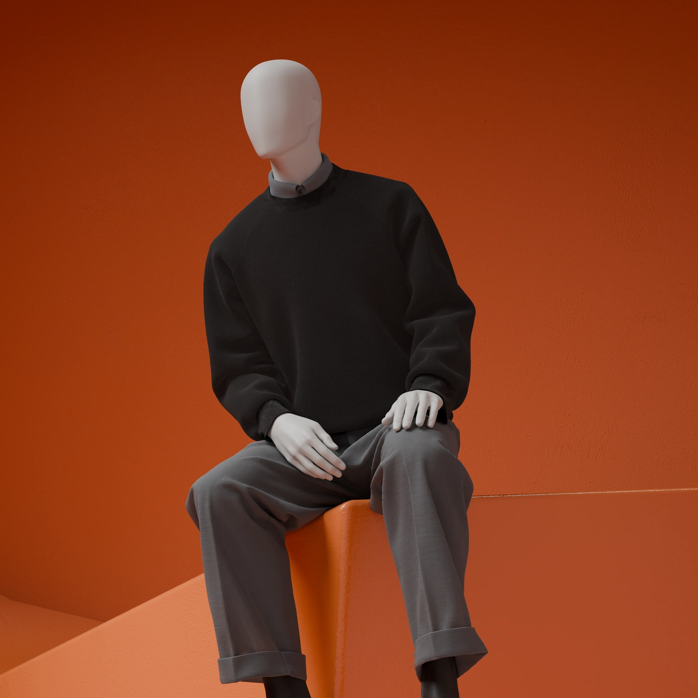 Seated male mannequin in a black knit sweater and grey cuffed trousers on an orange block against a rust-orange background